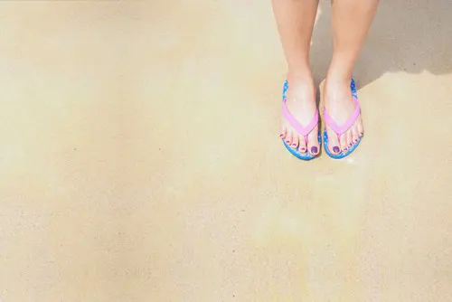 Feet wearing slippahs whilst standing on a sunny, sandy beach